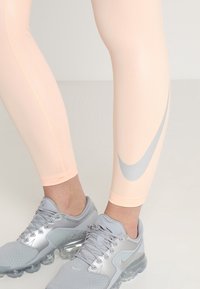 Jasno brzoskwiniowe legginsy sportowe z srebrnym logo Nike, o gładkiej strukturze i dopasowanym kroju, połączone z szarymi butami.