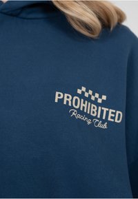 Sweat à capuche bleu marine en tissu doux présentant un logo brodé crème avec des accents à carreaux indiquant "PROHIBITED Racing Club".