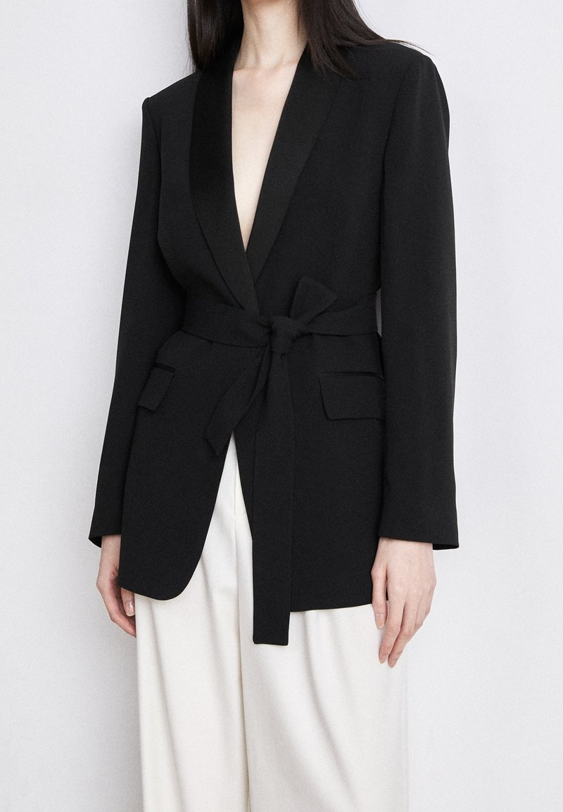 Femme portant un blazer noir ceinturé avec poches à rabat sur un pantalon blanc, debout devant un fond clair uni.
