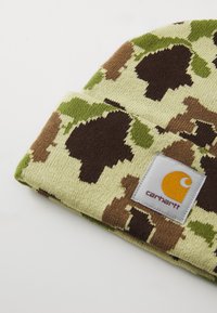 Căciulă tricotată de culoare verde deschis, cu un model de camuflaj în nuanțe de maro și măslin. Prezintă o eticheta pătrată cu text galben și negru. Textură moale.