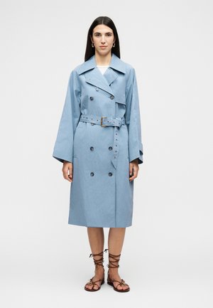 Kvinde med langt mørkt hår iført en lyseblå dobbeltradet trenchcoat med bælte og brune snøresandaler, stående mod en ensfarvet baggrund.