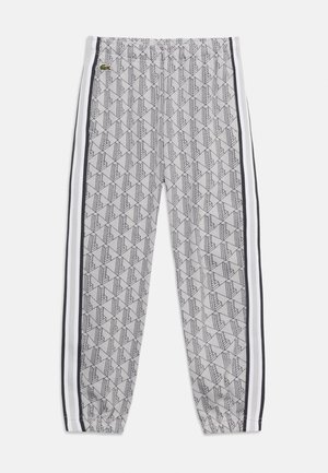 Grå joggingbukser med geometrisk sort mønster, sorte og hvide side striber, elastisk talje og ankellukninger samt grøn Lacoste-logo nær taljen.