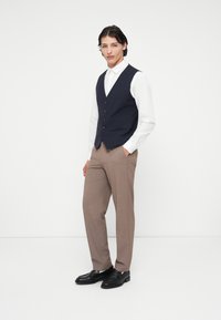 Colete azul-marinho com cinco botões sobre uma camisa branca, combinado com calças castanhas claras e sapatos pretos, apresentando um corte ajustado e uma textura suave.