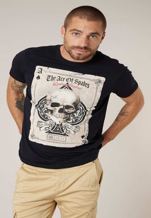 Homme aux cheveux courts et barbe, portant un t-shirt noir avec un grand graphique de crâne d'As de Pique et un pantalon cargo beige, les mains derrière le dos.