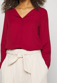 Blusa rossa con scollatura a V e una texture liscia, abbinata a pantaloni plissettati color crema a vita alta con cintura annodata.