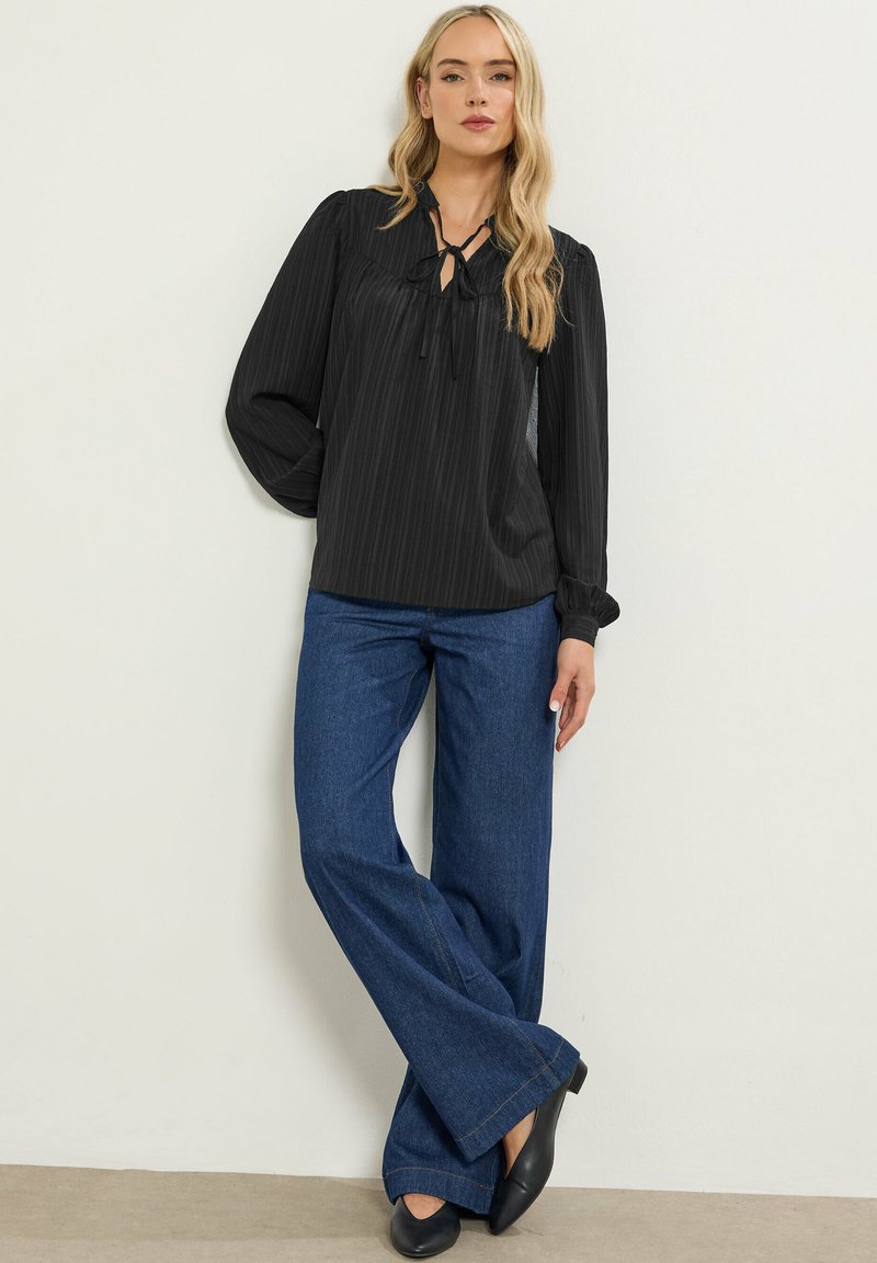 En blond kvinde iført en sort bluse med lange ærmer og bindebånd, blå bredbenede jeans og sorte spidse sko, stående mod en ensfarvet væg.