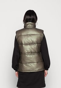 Kobieta z ciemnymi włosami, ubrana w metaliczny oliwkowozielony kamizelkę puchową na czarną sukienkę z długim rękawem, widziana z tyłu na jednolitym tle.