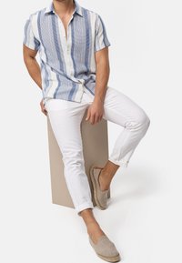 Camisa de botones de manga corta con rayas azules sobre tela crema, combinada con pantalones blancos y espadrilles beige sobre un pedestal neutro.
