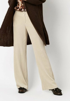 Breit geschnittene Cordhosen in Cremefarbe mit vertikalen Rippen, kombiniert mit einer braunen Strickjacke und schwarzen verzierten Schuhen.