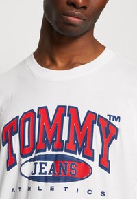 Tricou din bumbac alb cu un imprimeu grafic îndrăzneț în roșu și albastru "TOMMY JEANS". Mâneci scurte și decolteu rotund cu textură netedă.