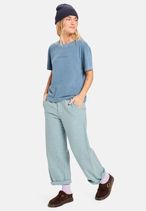 Blauwe katoenen t-shirt met korte mouwen, gecombineerd met lichtgroene geruite broek. Model draagt een marineblauwe beanie, paarse sokken en bruine klompen.
