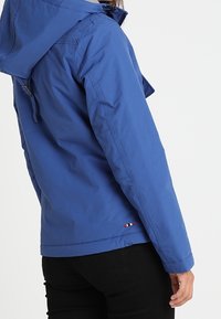 Osoba v modré bundě s kapucí s malým detaily trojbarevné vlajky u spodního lemu, v černých kalhotách, zobrazena zezadu.