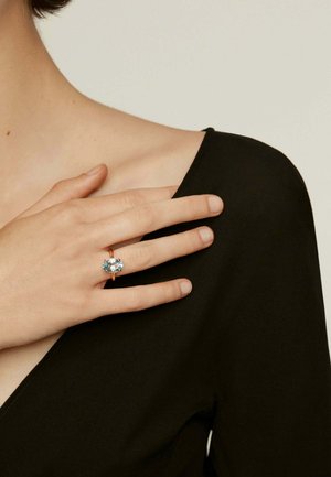 Anillo de oro rosa con una gran piedra preciosa azul en forma ovalada, llevado en una mano con un color de uñas neutro. El fondo está suavemente desenfocado.