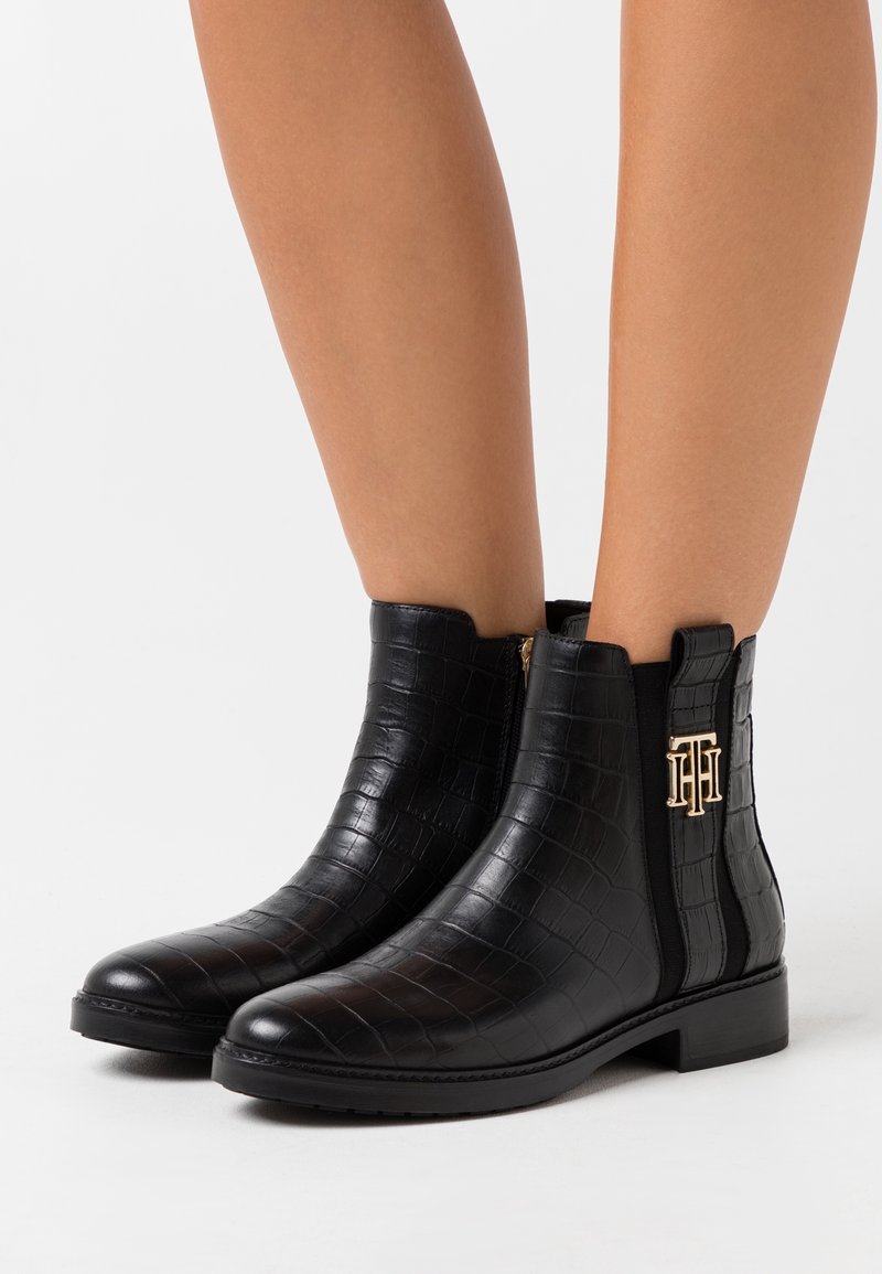 Bottes noires à cheville avec texture crocodile, fermeture éclair latérale, panneau élastique et logo "TH" en métal doré portées aux pieds d'une personne.