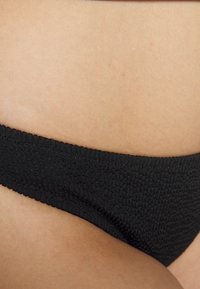 Tanga preta texturizada com um padrão trançado e uma cintura lisa, destacando um ajuste justo na área de pele exposta.