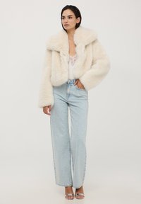 Giacca corta in finta pelliccia color crema con colletto ampio, indossata sopra un top bianco con pizzo e jeans azzurri a vita alta. Tacchi argentati.