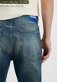 Blue denim jeans feature a straight cut, orange stitching, and a leather tag with "HUGO." The back pockets have a distinct shape and texture.