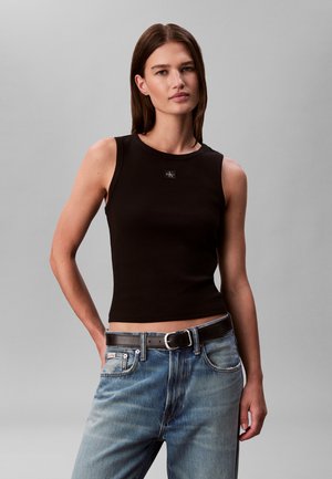 Kvinde med glat brunt hår iført en sort ærmeløs top og blå jeans med sort bælte mod en ensfarvet grå baggrund.