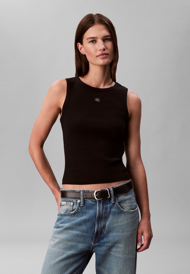 Kvinde med glat brunt hår iført en sort ærmeløs top og blå jeans med sort bælte mod en ensfarvet grå baggrund.