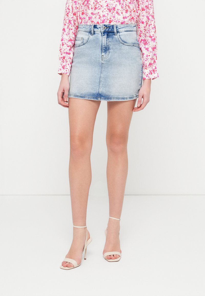 Φούστα μίνι από ανοιχτό μπλε τζιν με ψηλή μέση, τσέπες μπροστά και ξεβάμματα. Φορεμένη με γόβες σε τόνο του γυμνού με λουράκι στον αστράγαλο.