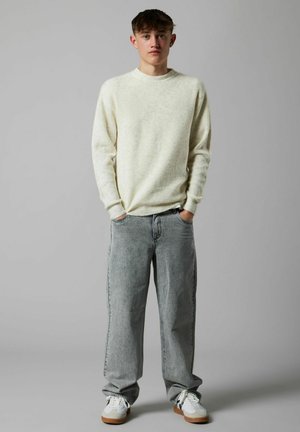 Giovane uomo in piedi con le mani nelle tasche, indossa un maglione lavorato a maglia color crema, jeans grigi larghi e sneakers bianche su uno sfondo grigio uniforme.