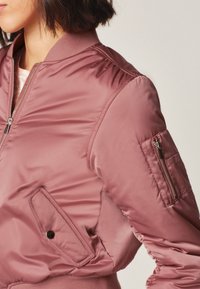 Veste bomber rose en nylon lisse avec poignets côtelés, fermeture éclair à l'avant et poches latérales. Comprend une poche supplémentaire zippée sur la manche.