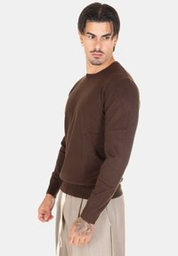 Maglione marrone a maniche lunghe con scollo rotondo, realizzato in tessuto morbido con un design aderente e bordo a costine. Tatuaggi visibili sulla mano sinistra.