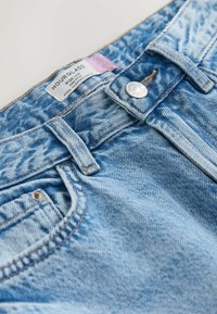 Blugi din denim albastru deschis cu talie înaltă și croială largă, prevăzuți cu un nasture argintiu și o etichetă pe care scrie „HOURGLASS”. Textură uzată vizibilă.