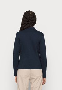 Osoba s dlouhými copy má na sobě těsně padnoucí námořnický blazer a béžové kalhoty, stojí zády k fotoaparátu proti jednoduchému světlem pozadí.