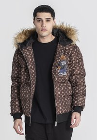 Giubbotto puffa marrone con cappuccio foderato in pelliccia, un motivo di logo e una toppa con orso sulla parte anteriore. Chiusura con zip e polsini a costine.