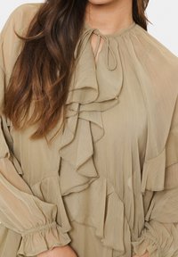 Beige blus med volangdetaljer, lättviktsmaterial, snörning i halsen och samlade ärmslut. Mjuk textur med en flödande design.