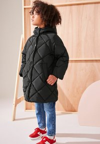 Geacă puffer neagră cu umplutură, cu glugă, având un model în diamond, buzunare laterale și o croială lejeră, purtată peste blugi albaștri și adidași roșii.