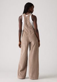 Brown corduroy overall with wide legs and large back pockets, featuring adjustable straps and a textured finish.