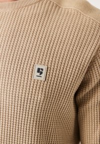 Pull beige en texture gaufrée avec un col côtelé. Présente une petite étiquette logo rectangulaire sur le coin supérieur gauche. Tissu doux, design près du corps.
