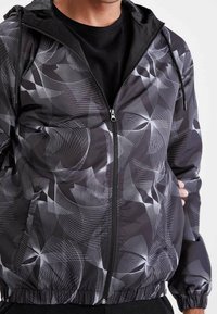 Sort letvægtsjakke med hvide abstrakte linjemønstre, lynlås foran, elastiske manchetter og kant, med hætte og sidelommer.