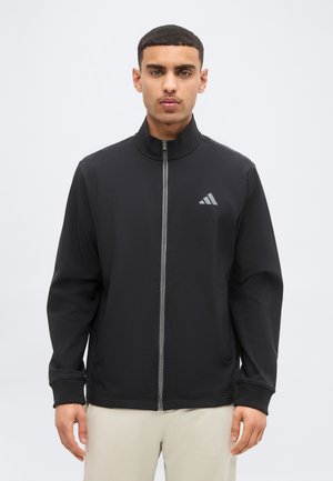 Férfi fekete cipzáras sportdzsekit viselve, rajta az Adidas logó a mellén, egyszínű világos háttér előtt áll.