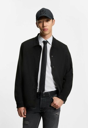 Junger Mann mit schwarzer Hugo-Kappe, schwarzer Jacke über weißem gestreiftem Hemd, schwarzer Krawatte und abgenutzten schwarzen Jeans, stehend vor weißem Hintergrund.