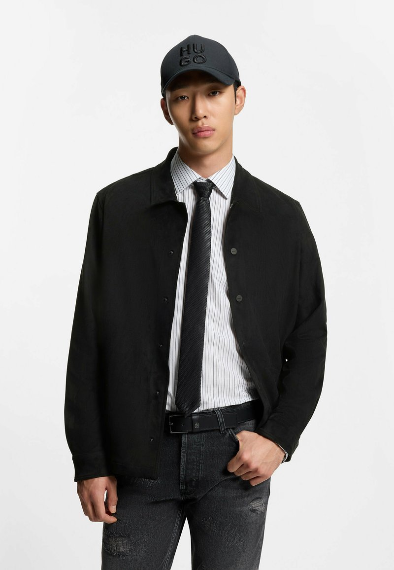 Joven con gorra negra de Hugo, chaqueta negra sobre camisa blanca a rayas, corbata negra y vaqueros negros desgastados, de pie frente a un fondo blanco.