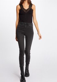 Czarny bodysuit z koronkowymi wykończeniami zestawiony z ciemnoszarymi spodniami skinny z wysokim stanem oraz czarnymi botkami za kostkę, prezentujący dopasowaną sylwetkę.