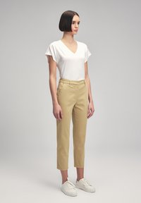 Top bianco a maniche corte con scollo a V e dettagli in pizzo, abbinato a pantaloni beige slim-fit e sneakers bianche, in piedi contro uno sfondo grigio.