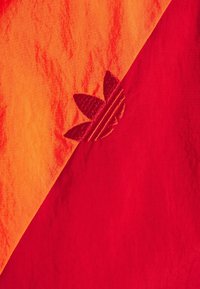 Orange och röd tyg med en broderad röd Adidas trefoil-logotyp vid det diagonala sömmen mellan de två färgerna.