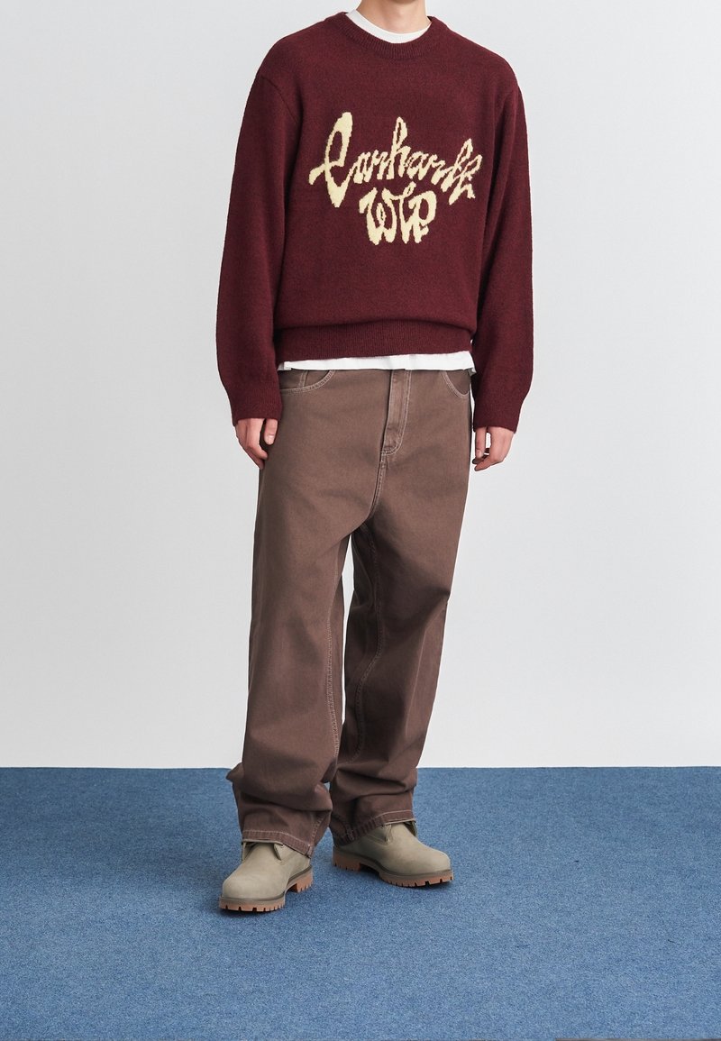 Personne portant un pull bordeaux avec texte, un pantalon marron ample, des bottes beiges, debout sur un tapis bleu contre un mur gris.