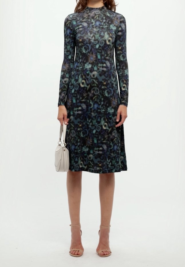 Femme portant une robe à manches longues, longueur genoux, avec un motif abstrait sombre, tenant un petit sac à main blanc et portant des talons couleur chair.