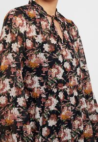 Gros plan sur un blouse noire transparente avec un motif floral multicolore et un décolleté keyhole noué avec un fin nœud.