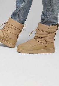 Brune sneboots med en tekstureret stofoverdel, elastiske snørebånd og gummisåler. Bemærkelsesværdige detaljer inkluderer et broderet design og logo på siden.