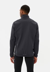 Giacca softshell grigio scuro con collo alto, tasche con zip e dettagli riflettenti. Texture liscia e design aderente per un uso attivo.