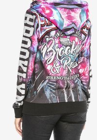 Hoodie med et levende design i pink, blå og sort, der har ordene "Brooklyn & Rock", vinger, kraniet grafik, og teksten "Styrke & Mystik" på ryggen.