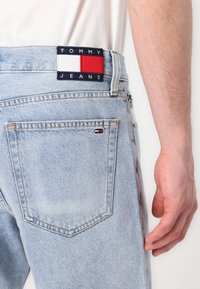 Light blue denim jeans with a rectangular navy and colored logo patch at the waistband and a small logo detail on the back pocket.