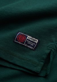 Camiseta de algodón verde con una etiqueta azul marino que presenta "SUPERDRY" y "VINTAGE ATHLETIC OUTFITTERS" en texto blanco y acentos rojos.