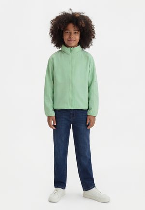 Kind mit lockigem Haar steht in einer hellgrünen Reißverschlussjacke, dunkelblauen Jeans und weißen Turnschuhen vor einem einfarbigen hellen Hintergrund.